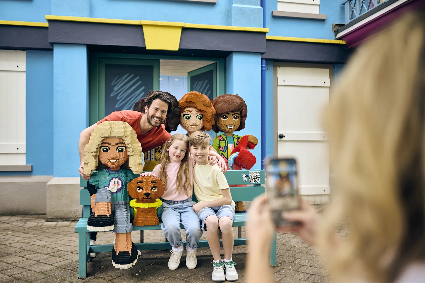 Father, Son And Daughter On Bench With LEGO Friends Models smiling at Mum in foreground taking a picture on her phone