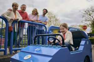 Young girl driving a LEGO City Car with her family smiling over at her in the background behind blue barriers
