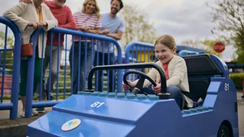 Young girl driving a LEGO City Car with her family smiling over at her in the background behind blue barriers